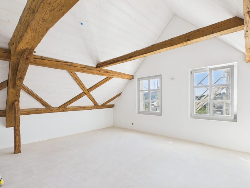 Heller Dachraum einer 4.5-Zimmer-Attikawohnung in der «Gelben Fabrik» mit sichtbaren Holzbalken, weisser Decke und grossen Fenstern.