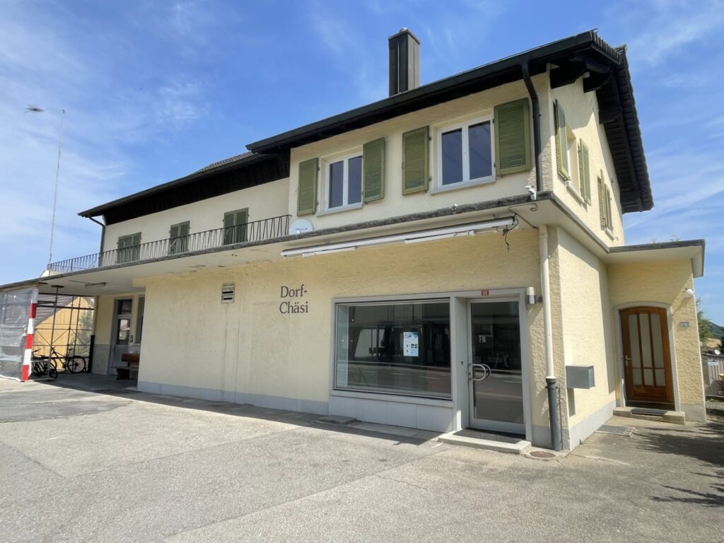 Gebäude der Dorf-Chäsi, alte Käsi, Rickenbach mit heller Fassade, grünen Fensterläden und Balkon im Obergeschoss.
