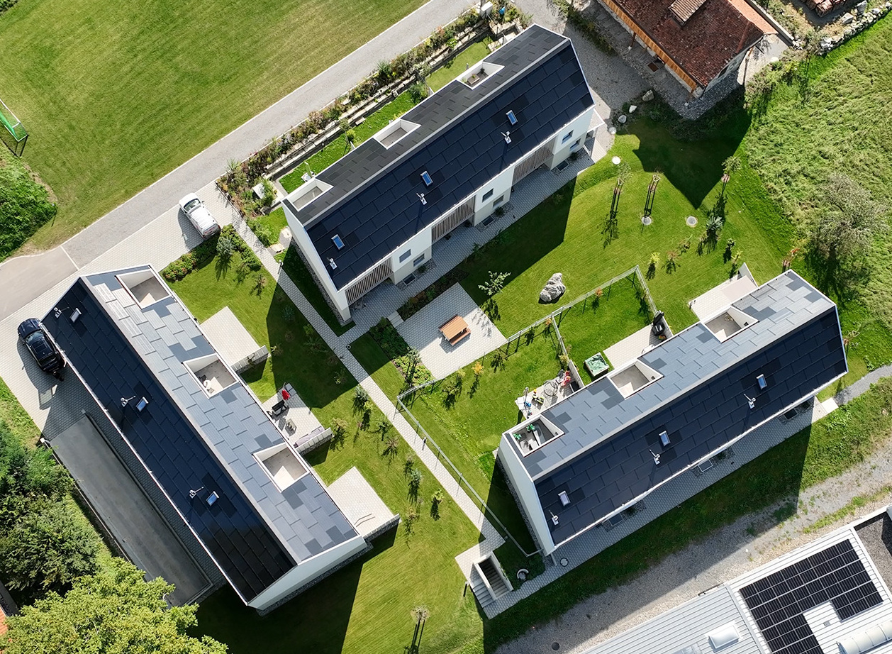 Luftaufnahme einer Neubauüberbauung mit modernen Reiheneinfamilienhäusern an der Schmiedgasse in Gontenschwil. Drei längliche Gebäudekörper mit dunklen Dächern und Solaranlagen umschließen eine begrünte Gartenanlage mit Wegen, Sitzplätzen und Spielbereichen.